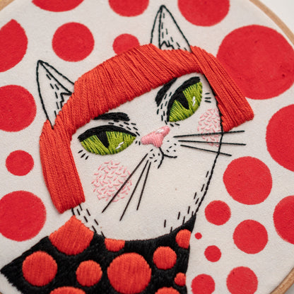 May include: Embroidered white cat with red hair and green eyes, wearing a black and red polka dot shirt. The background is white with red polka dots.