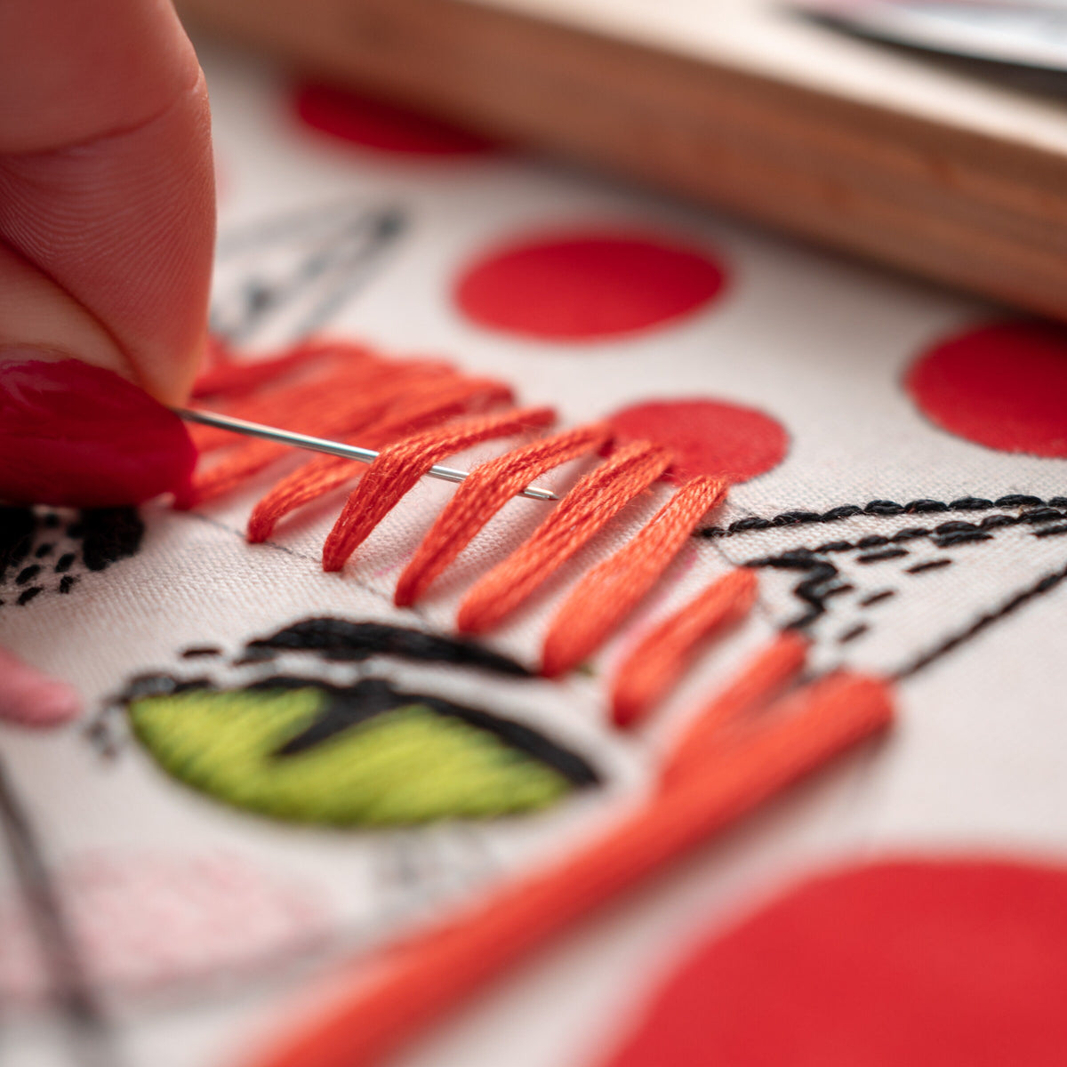 May include: Close-up of a person's hand embroidering a red and white design on a white fabric. The design includes a green eye and orange stitching.