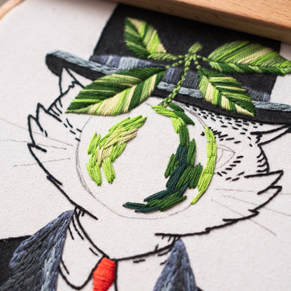 May include: Embroidery art of a cat wearing a top hat and suit, with a red tie. The cat's eyes are embroidered with green thread, and a green leaf design is above the cat's head. The embroidery is on a white fabric.