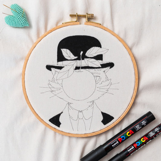 May include: An embroidery hoop with a cat design wearing a bowler hat and holding an apple, inspired by Magritte's art. The design is in black on a white fabric. Two black Uni Posca markers are next to the hoop.