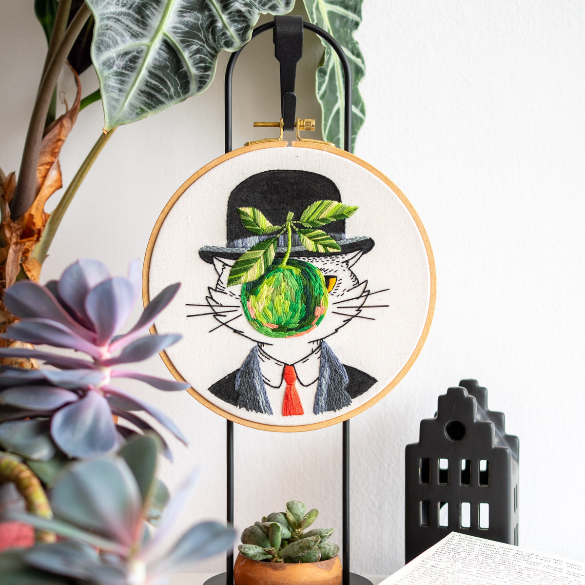 May include: Embroidered artwork in a wooden hoop features a cat in a bowler hat, suit, and red tie, with a green apple in place of its face. The cat is set against a white background, mounted on a black metal stand.