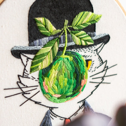 May include: Embroidered artwork featuring a cat wearing a black bowler hat and a suit. The cat is holding a green, round fruit with a leaf in its mouth. The embroidery uses various shades of green, yellow, and black thread on a white background.