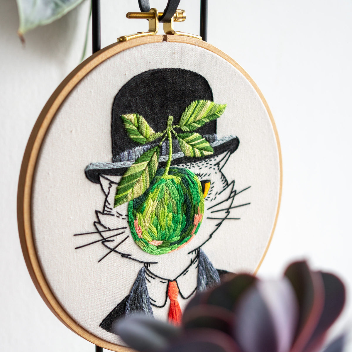 May include: Embroidered artwork in a wooden hoop features a cat in a bowler hat with a green apple and leaves. The cat wears a suit and tie. The embroidery is on a white background with a black ribbon and gold hardware for hanging.