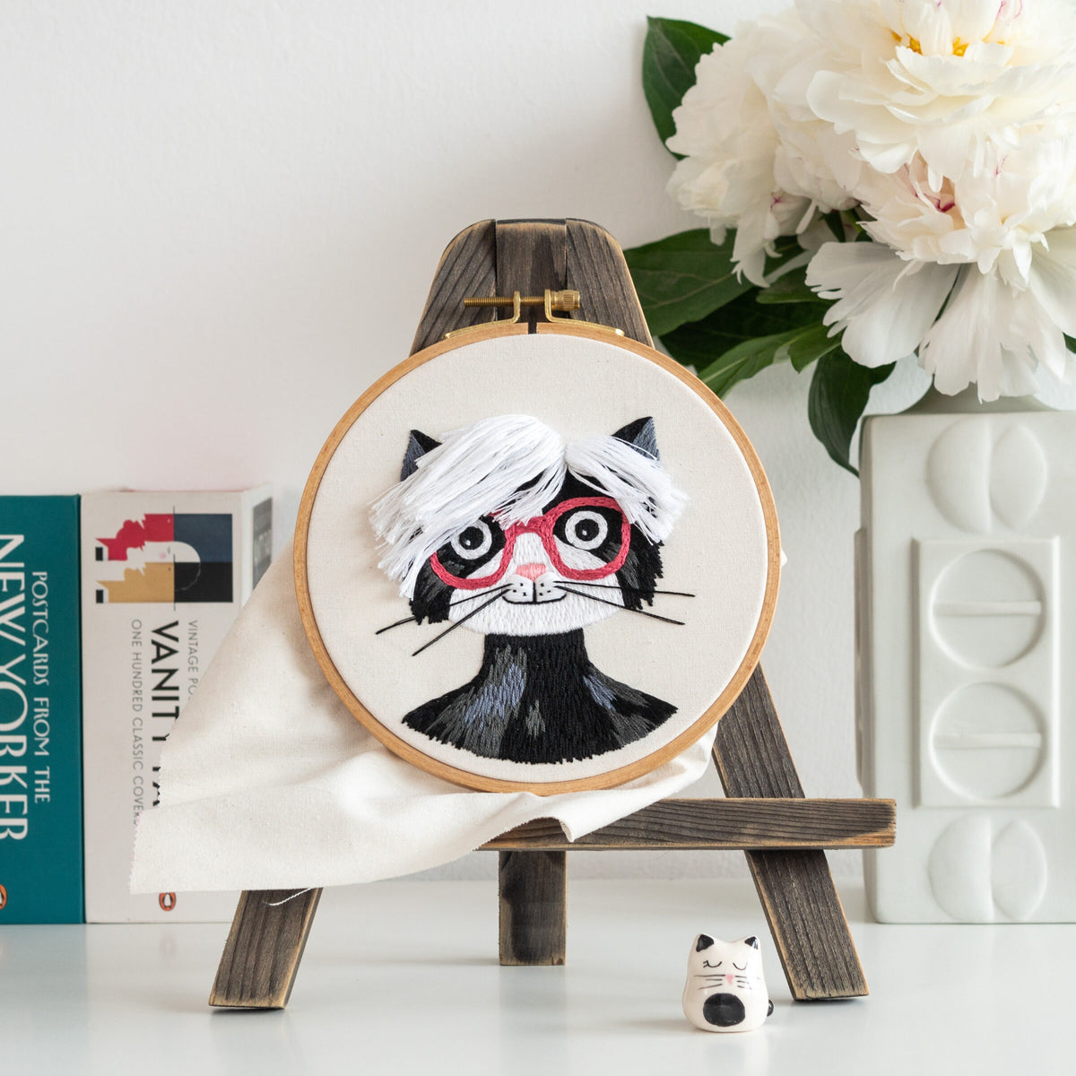 May include: Embroidered hoop art featuring a black cat with white hair and red glasses. The cat is wearing a black turtleneck. The hoop is on a wooden easel.
