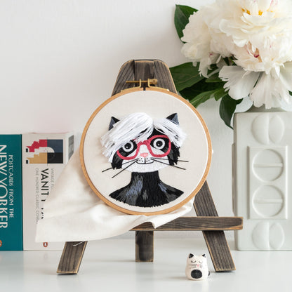 May include: Embroidered hoop art featuring a black cat with white hair and red glasses. The cat is wearing a black turtleneck. The hoop is on a wooden easel.