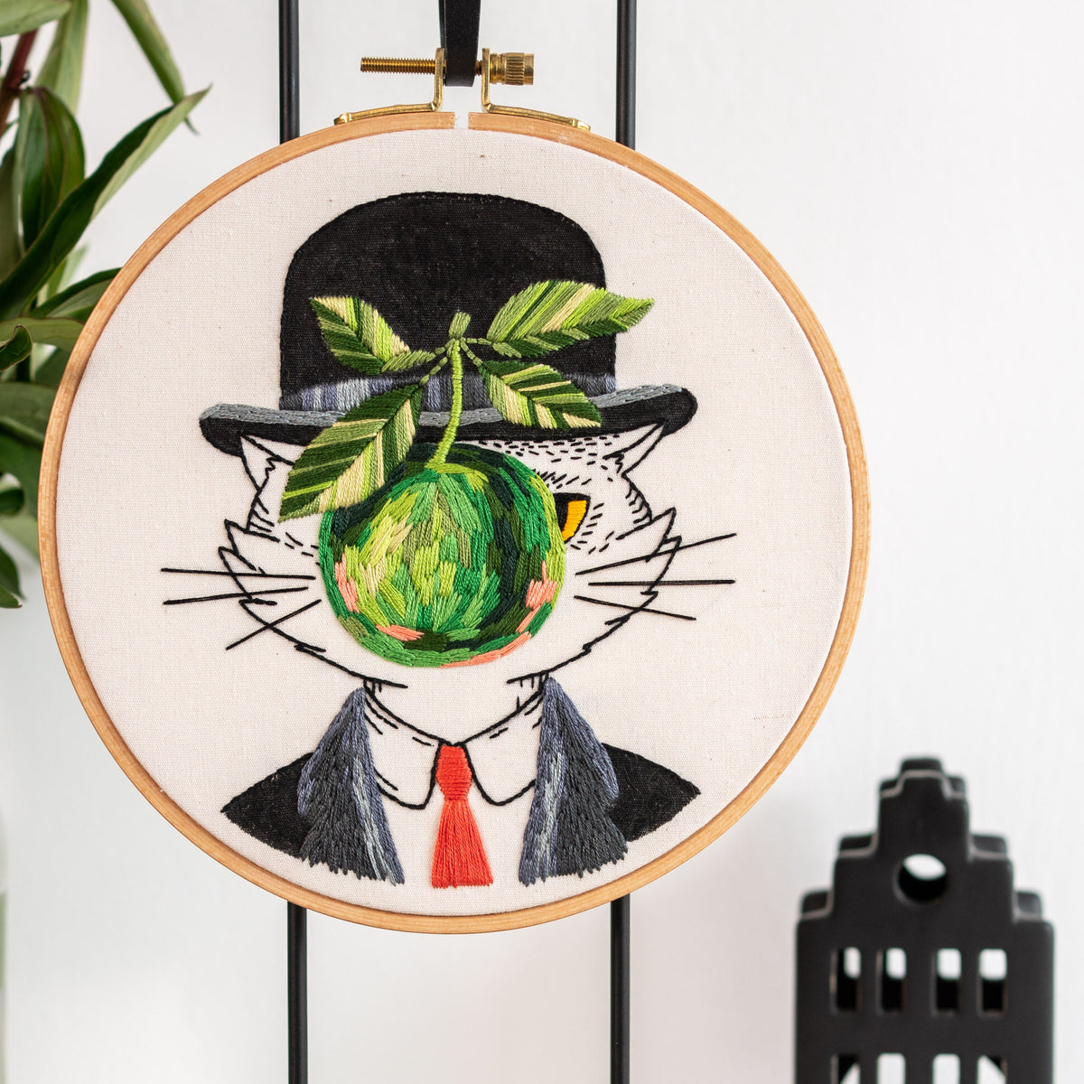May include: Embroidered hoop art featuring a white cat wearing a black top hat with a green apple covering its face. The cat is wearing a gray suit with a red tie.