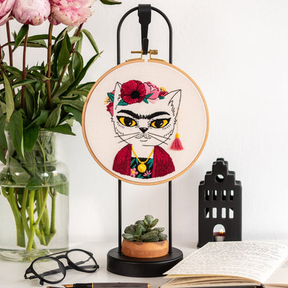 May include: Embroidered hoop art featuring a white cat with a red and pink flower crown, wearing a red jacket and a gold necklace. The hoop is on a black stand with a small potted plant.