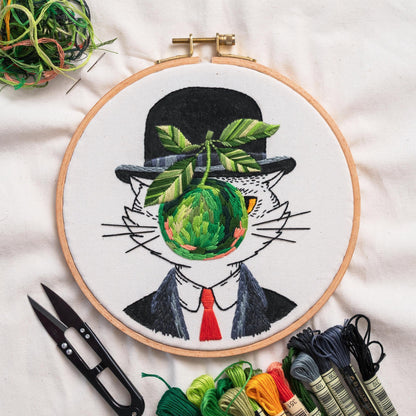 May include: An embroidery hoop featuring a cat in a bowler hat, suit, and red tie, with a green apple in place of a face. The apple and leaves are intricately stitched. Embroidery floss and scissors are visible.