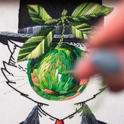 May include: Close-up of an embroidery project featuring a cat wearing a hat and suit. The cat's eye is a green apple with leaves. The embroidery uses various shades of green, orange, and black thread on a white background. A hand is holding a needle