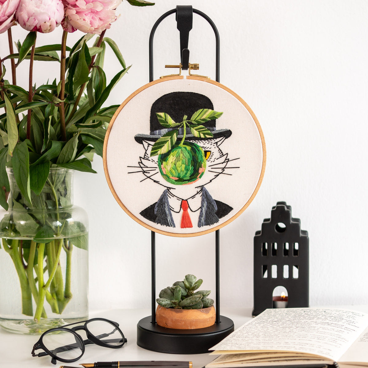 May include: Embroidered hoop art featuring a black and white cat wearing a black top hat and holding a green apple in its mouth. The cat is surrounded by a white background and the hoop is hanging from a black stand.
