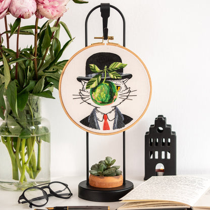 May include: Embroidered hoop art featuring a black and white cat wearing a black top hat and holding a green apple in its mouth. The cat is surrounded by a white background and the hoop is hanging from a black stand.