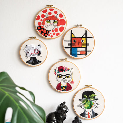 May include: Five embroidery hoops with different cat designs. The hoops are white with black outlines and feature colorful embroidery. The cats are all different styles, including a cat with a red and white polka dot background, a cat with a geometr