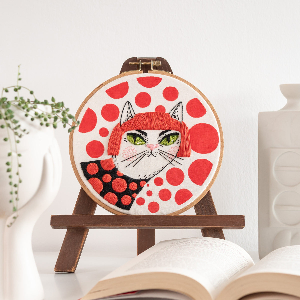 May include: Embroidered hoop art featuring a white cat with red hair wearing a black and red polka dot dress. The cat is set against a red and white polka dot background.