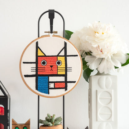 May include: A white embroidery hoop with a colorful geometric cat design. The cat is red, yellow, blue, and black. The hoop is on a black stand.