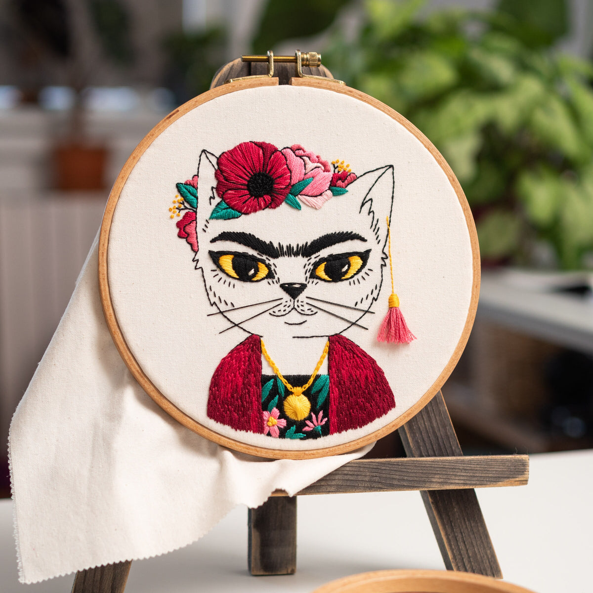 May include: Embroidered hoop art featuring a white cat with a red and black floral crown, wearing a red jacket and a gold necklace. The cat has a black mustache and eyebrows.