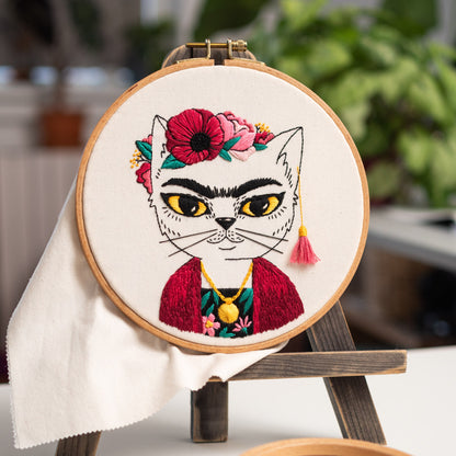 May include: Embroidered hoop art featuring a white cat with a red and black floral crown, wearing a red jacket and a gold necklace. The cat has a black mustache and eyebrows.