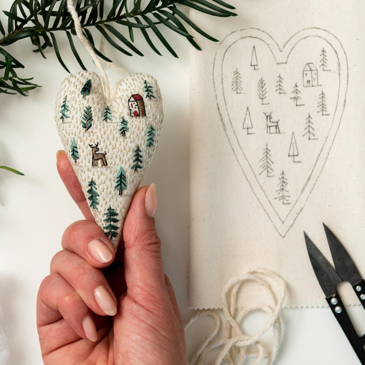 May include: A white heart-shaped fabric ornament with green embroidered trees and a red house. The ornament is being held by a hand. A white heart-shaped fabric with a black outline of trees and a house is in the background.