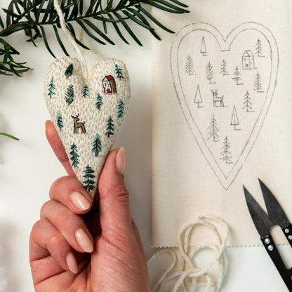 May include: A white heart-shaped fabric ornament with green embroidered trees and a red house. The ornament is being held by a hand. A white heart-shaped fabric with a black outline of trees and a house is in the background.