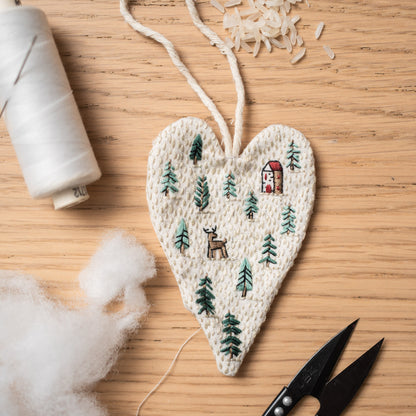 May include: A white heart-shaped fabric ornament with green embroidered trees, a small red house, and a deer. The ornament is on a wooden surface with white thread and a spool of white thread.