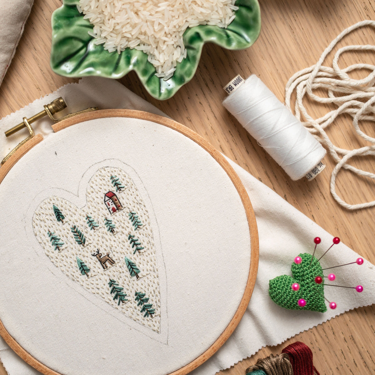 May include: A white embroidery hoop with a heart-shaped design of a forest scene with a small house and a deer. The hoop is on a white fabric surface with a spool of white thread, a green heart-shaped pincushion with pink pins, and a bowl of white r