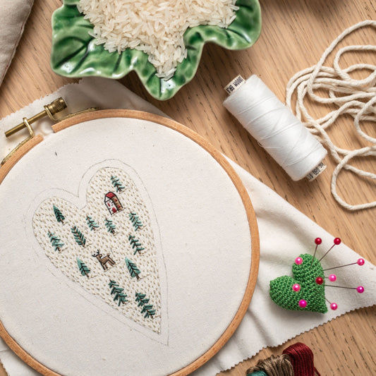 May include: A white embroidery hoop with a heart-shaped design of a forest scene with a small house and a deer. The hoop is on a white fabric surface with a spool of white thread, a green heart-shaped pincushion with pink pins, and a bowl of white r