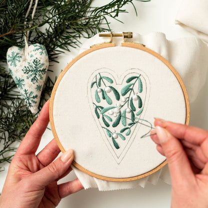 May include: A close-up of a hand embroidering a green mistletoe design in the shape of a heart on white fabric in a wooden embroidery hoop.
