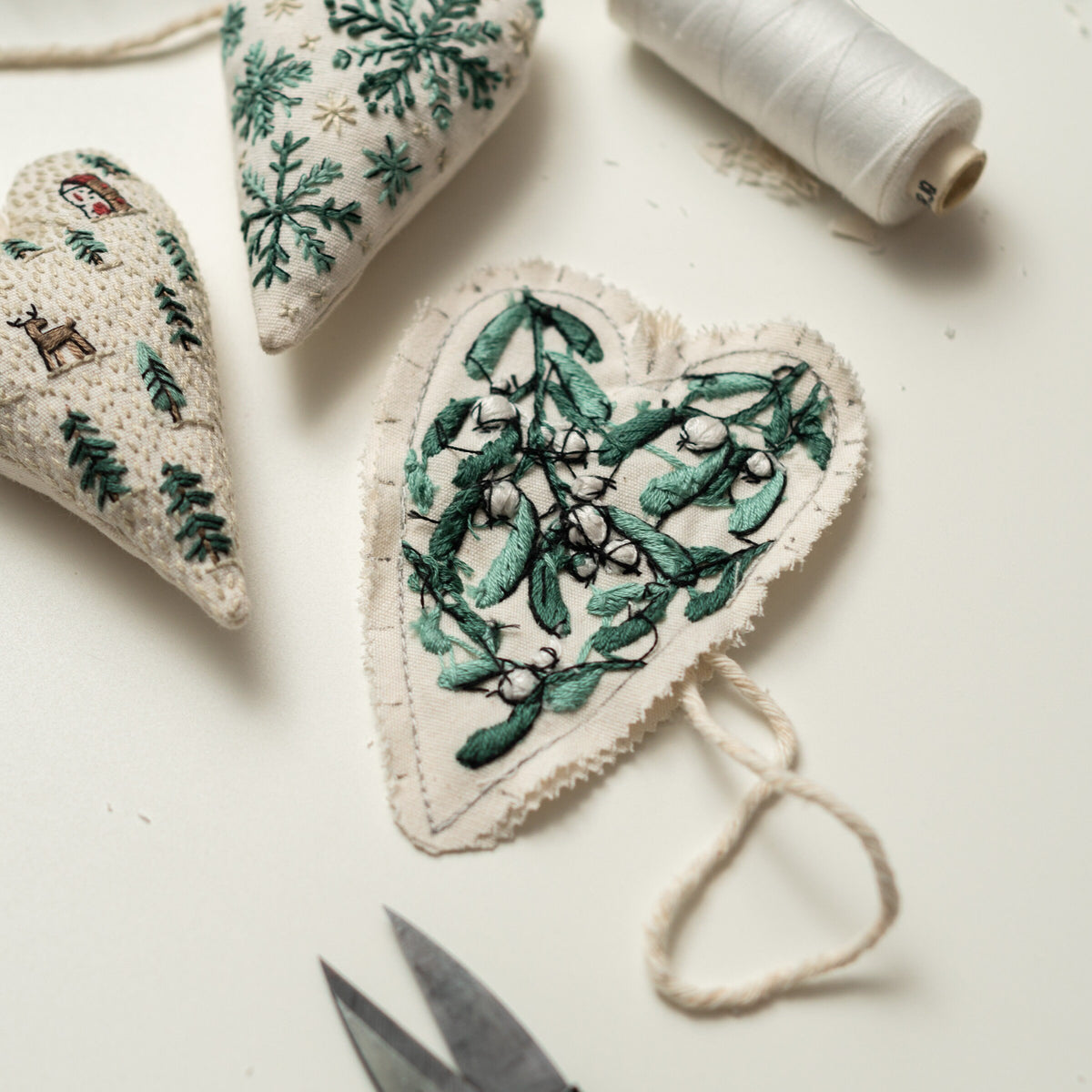May include: Three handmade felt heart ornaments with green embroidery. The ornaments feature a snowflake, a winter scene, and a sprig of mistletoe. The ornaments are hanging from a white string.