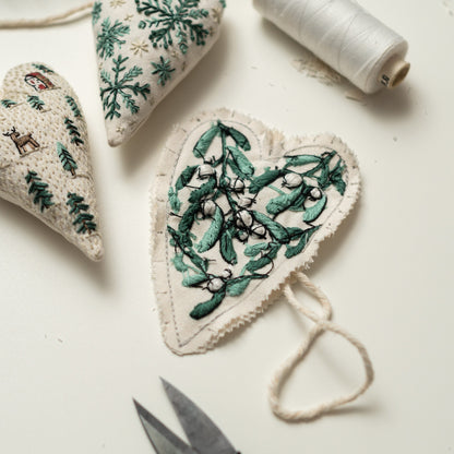 May include: Three handmade felt heart ornaments with green embroidery. The ornaments feature a snowflake, a winter scene, and a sprig of mistletoe. The ornaments are hanging from a white string.