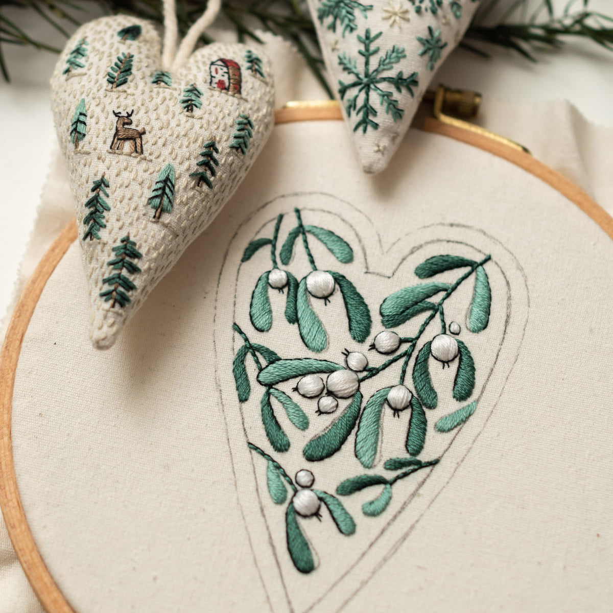 May include: A white embroidery hoop with a green and white embroidered heart-shaped design of mistletoe. The mistletoe is a traditional Christmas decoration.