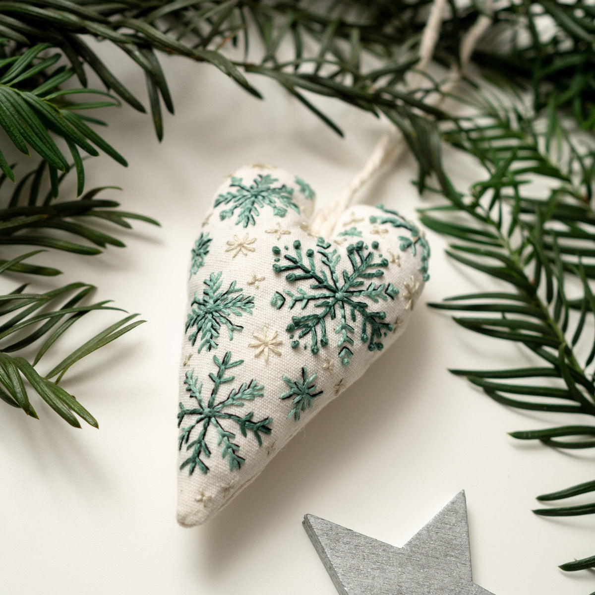 May include: A white heart-shaped ornament with green and white embroidered snowflakes. The ornament is hanging from a white string.