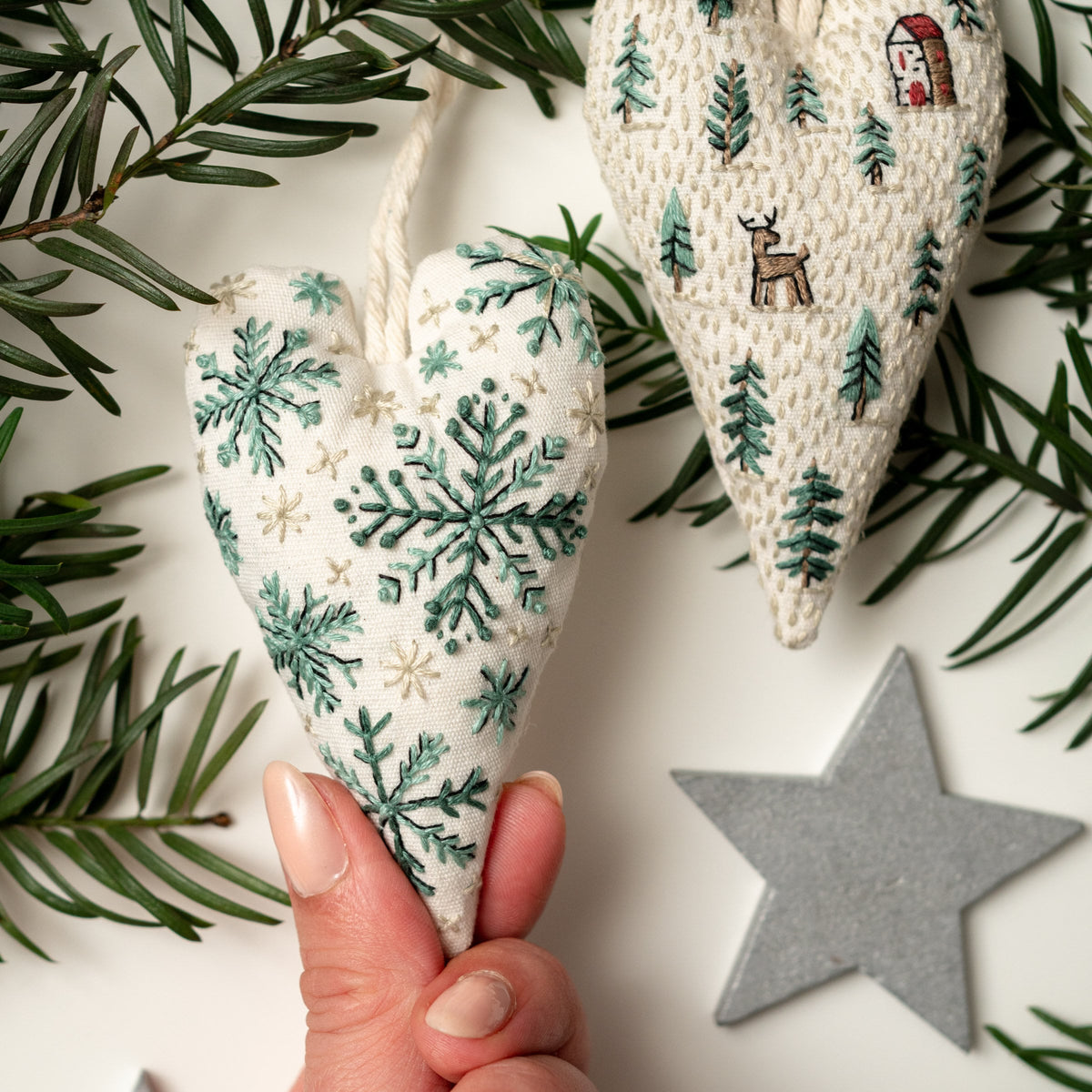 May include: Two white felt heart-shaped ornaments with green embroidery. One ornament features a snowflake design, the other features a winter scene with trees and a small house.