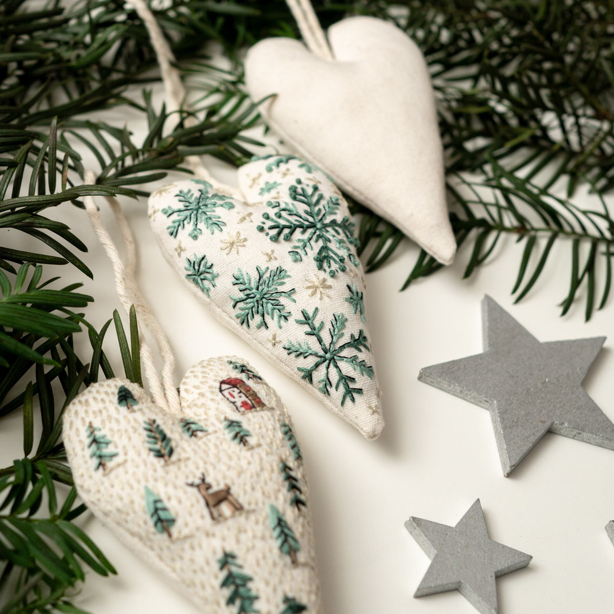 May include: Three white fabric heart ornaments with different embroidered designs. The ornaments are hanging from a green pine branch. The designs include snowflakes, pine trees, and a snowman.
