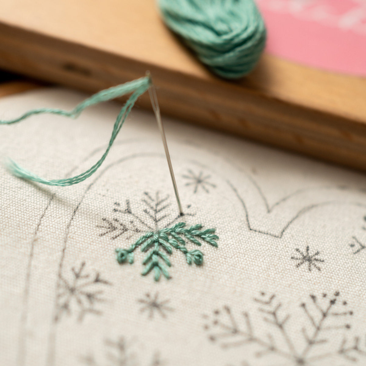 May include: Close-up of a needle and thread being used to embroider a green snowflake design on a white fabric. The fabric has a pattern of smaller snowflakes embroidered in black thread.
