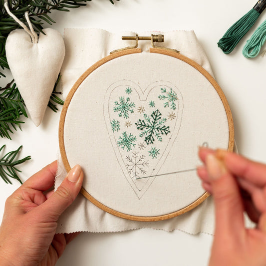 May include: A close-up of a hand embroidering a heart-shaped design with green and white snowflakes on a white fabric stretched in a wooden embroidery hoop.