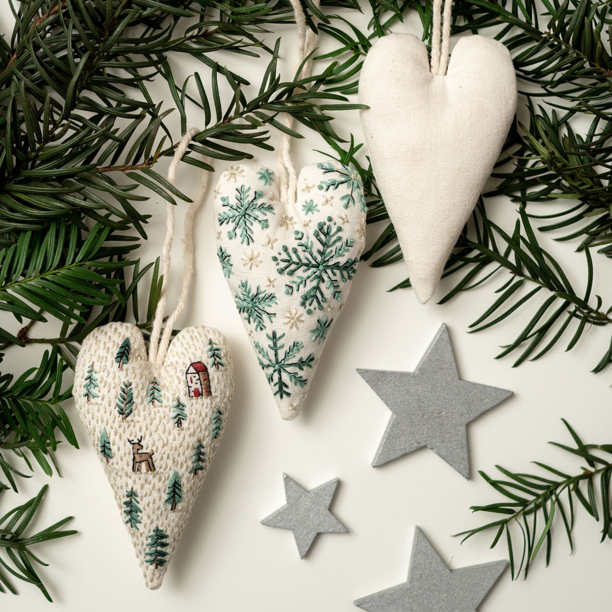 May include: Three handmade felt heart ornaments with different embroidered designs. One heart features a winter scene with snowflakes, another has a forest scene with trees and a deer, and the third is plain white. The ornaments are hanging from a w