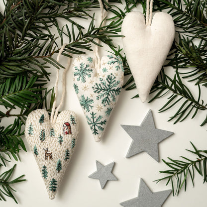 May include: Three handmade felt heart ornaments with different embroidered designs. One heart features a winter scene with snowflakes, another has a forest scene with trees and a deer, and the third is plain white. The ornaments are hanging from a w