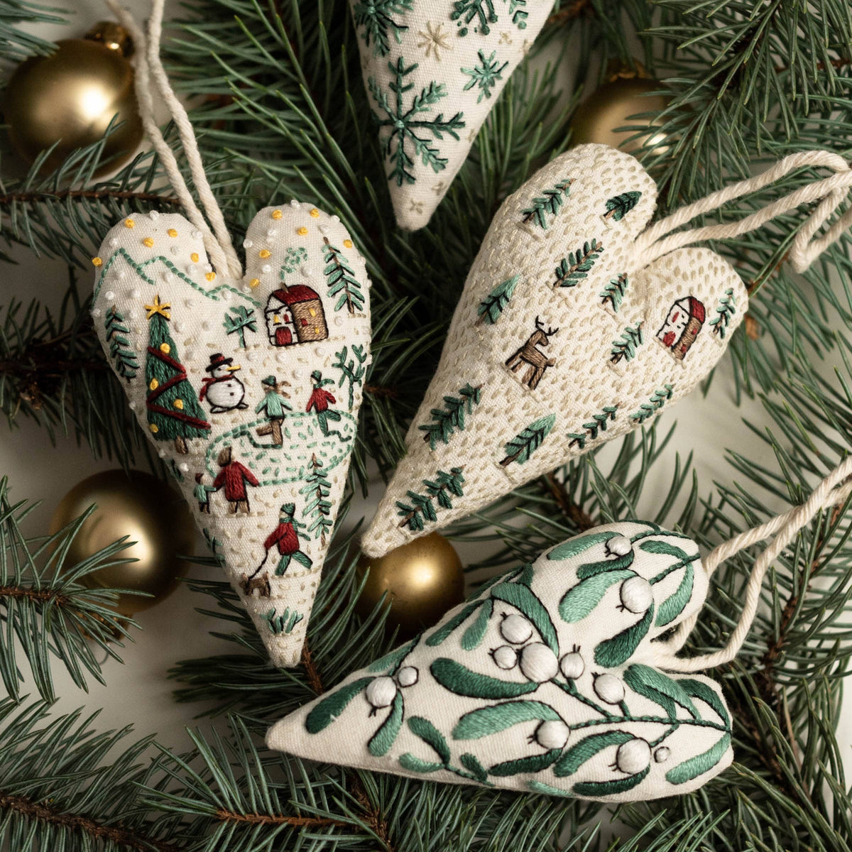May include: Four handmade heart-shaped ornaments with embroidered winter scenes. The ornaments are made of white fabric with green and gold embroidery. The scenes include a winter village, a snowy forest, and a sprig of mistletoe.