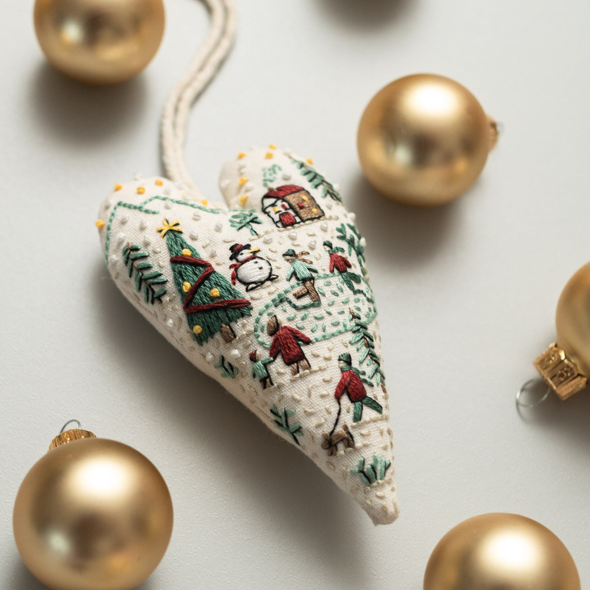 May include: A white heart-shaped ornament with a winter scene embroidered on it. The scene includes a snowman, a Christmas tree, and people walking in the snow. The ornament is surrounded by gold-colored ornaments.