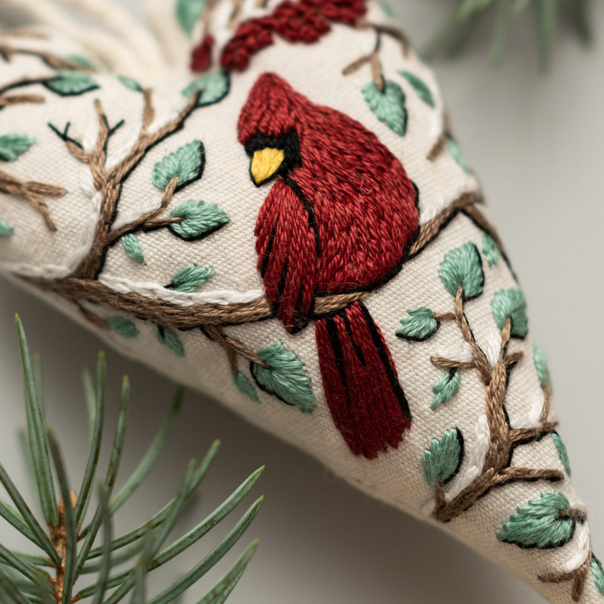 May include: A heart-shaped embroidered ornament with a red cardinal perched on a branch with green leaves. The ornament is made of white fabric and has a brown branch with white snow-like accents.