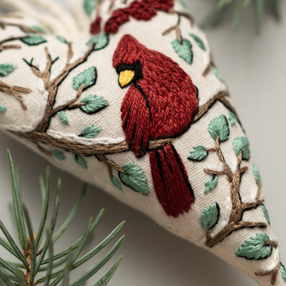 May include: A heart-shaped embroidered ornament with a red cardinal perched on a branch with green leaves. The ornament is made of white fabric and has a brown branch with white snow-like accents.