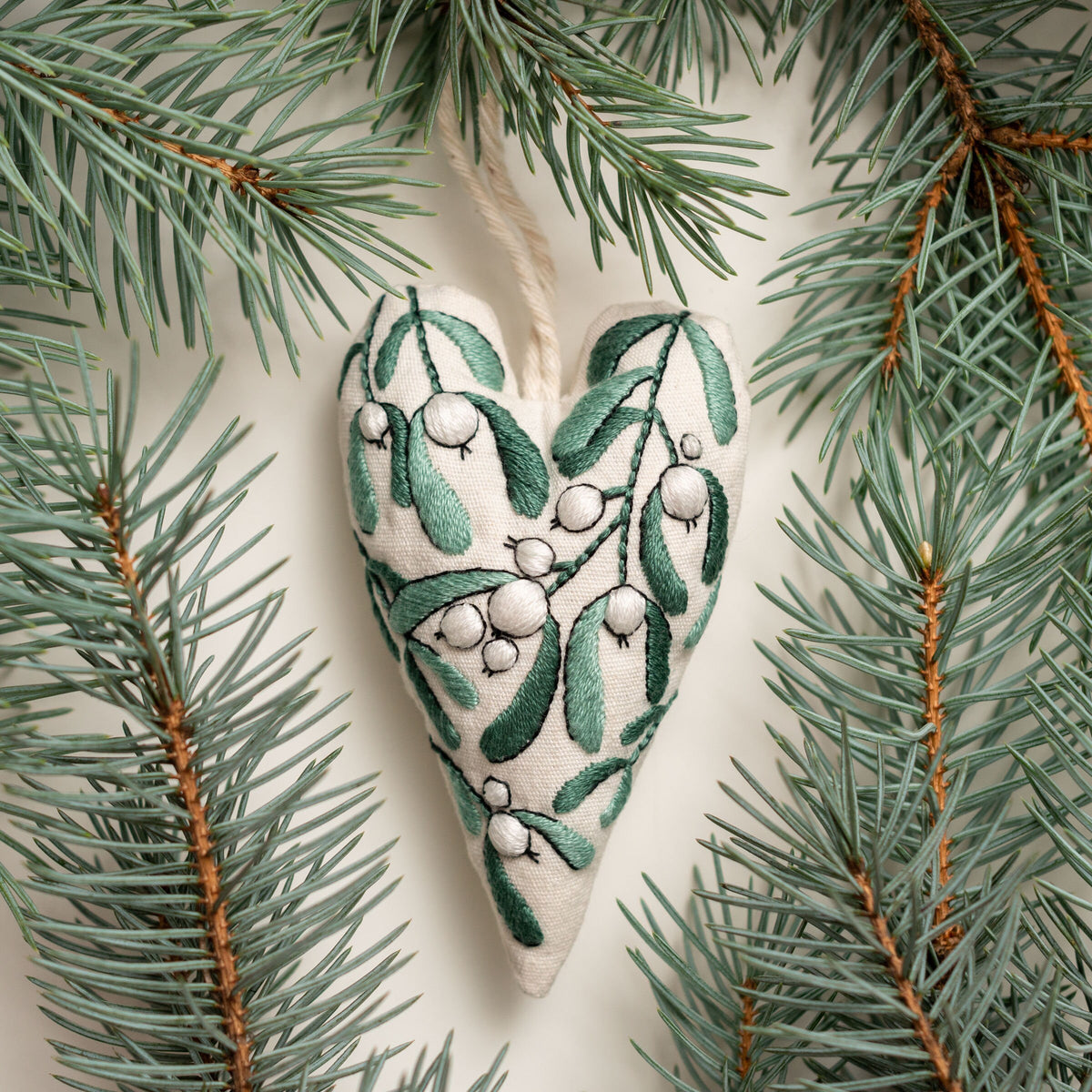 May include: A white heart-shaped ornament with green embroidery of mistletoe sprigs and white berries.