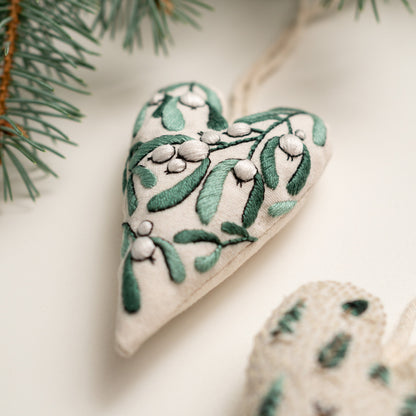 May include: A white heart-shaped ornament with green and white embroidery of mistletoe.