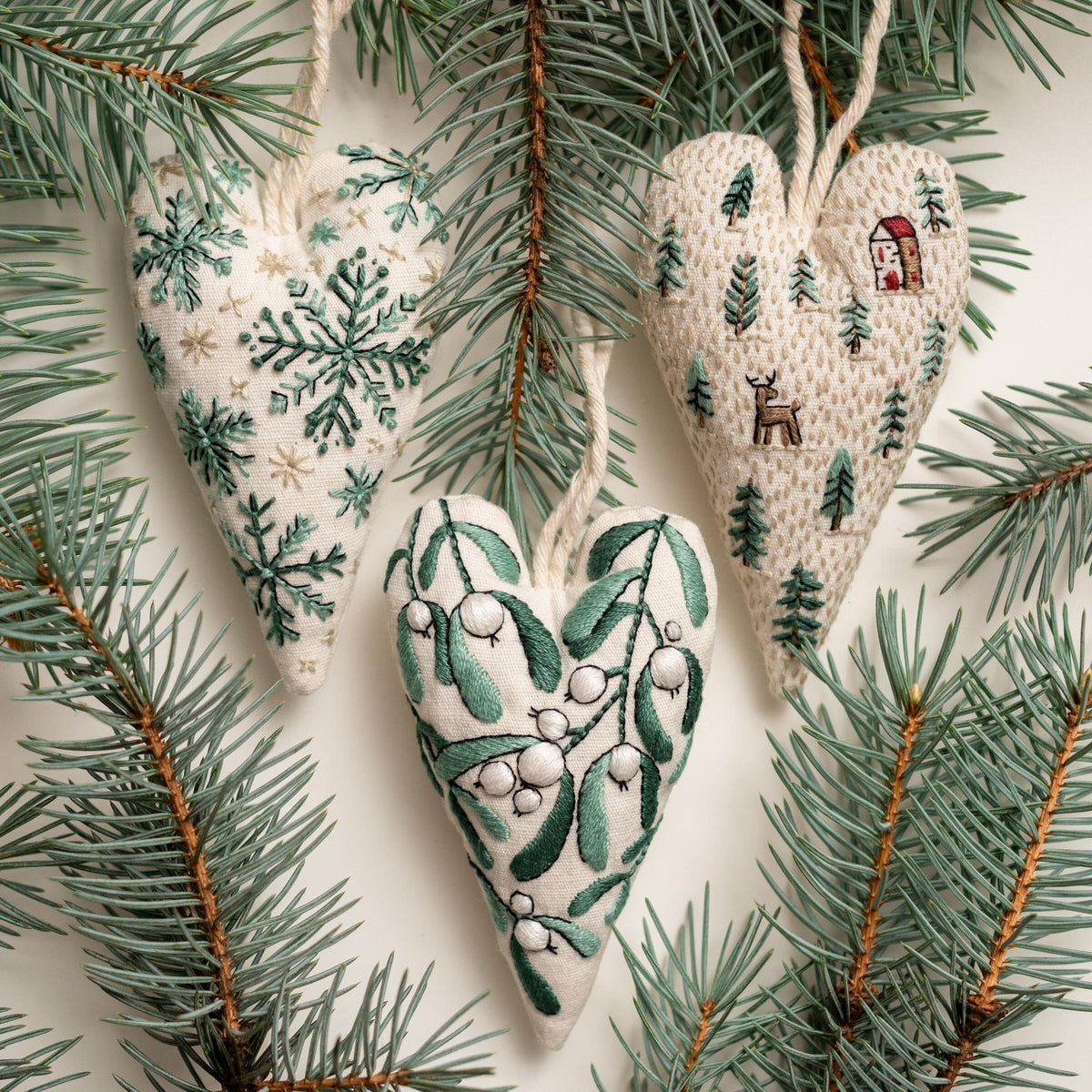 May include: Three handmade heart-shaped ornaments with different embroidered designs. The ornaments are made of white fabric with green embroidery. One ornament features a snowflake design, another features a winter scene with a house and a deer, an