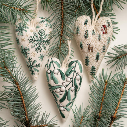 May include: Three handmade heart-shaped ornaments with different embroidered designs. The ornaments are made of white fabric with green embroidery. One ornament features a snowflake design, another features a winter scene with a house and a deer, an