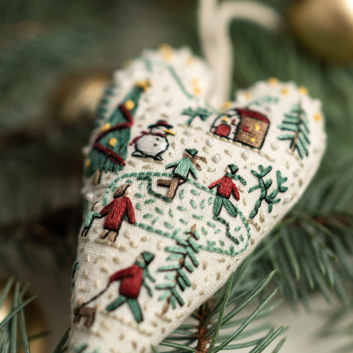 May include: A white heart-shaped ornament with embroidered winter scenes, including a snowman, people, trees, and a house. The ornament is decorated with small white beads.