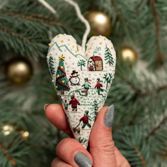 May include: A white heart-shaped ornament with embroidered details of a winter scene. The scene includes a snowman, a house, and people ice skating. The ornament is decorated with small white beads.