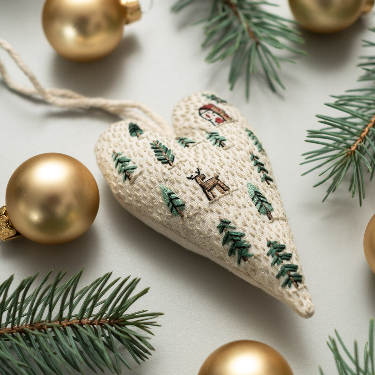 May include: A white heart-shaped ornament with green embroidered pine trees, a reindeer, and a small house. The ornament is hanging from a white string and is surrounded by gold ornaments and pine branches.