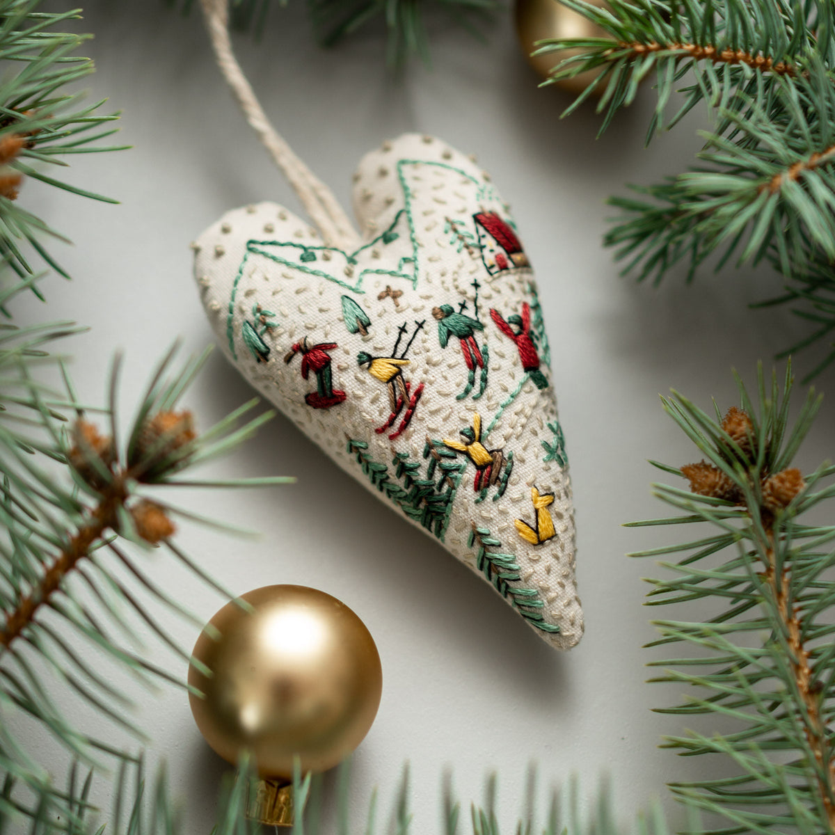 May include: A white heart-shaped ornament with a winter scene embroidered on it. The scene includes skiers, snowboarders, and trees. The ornament is hanging from a string.