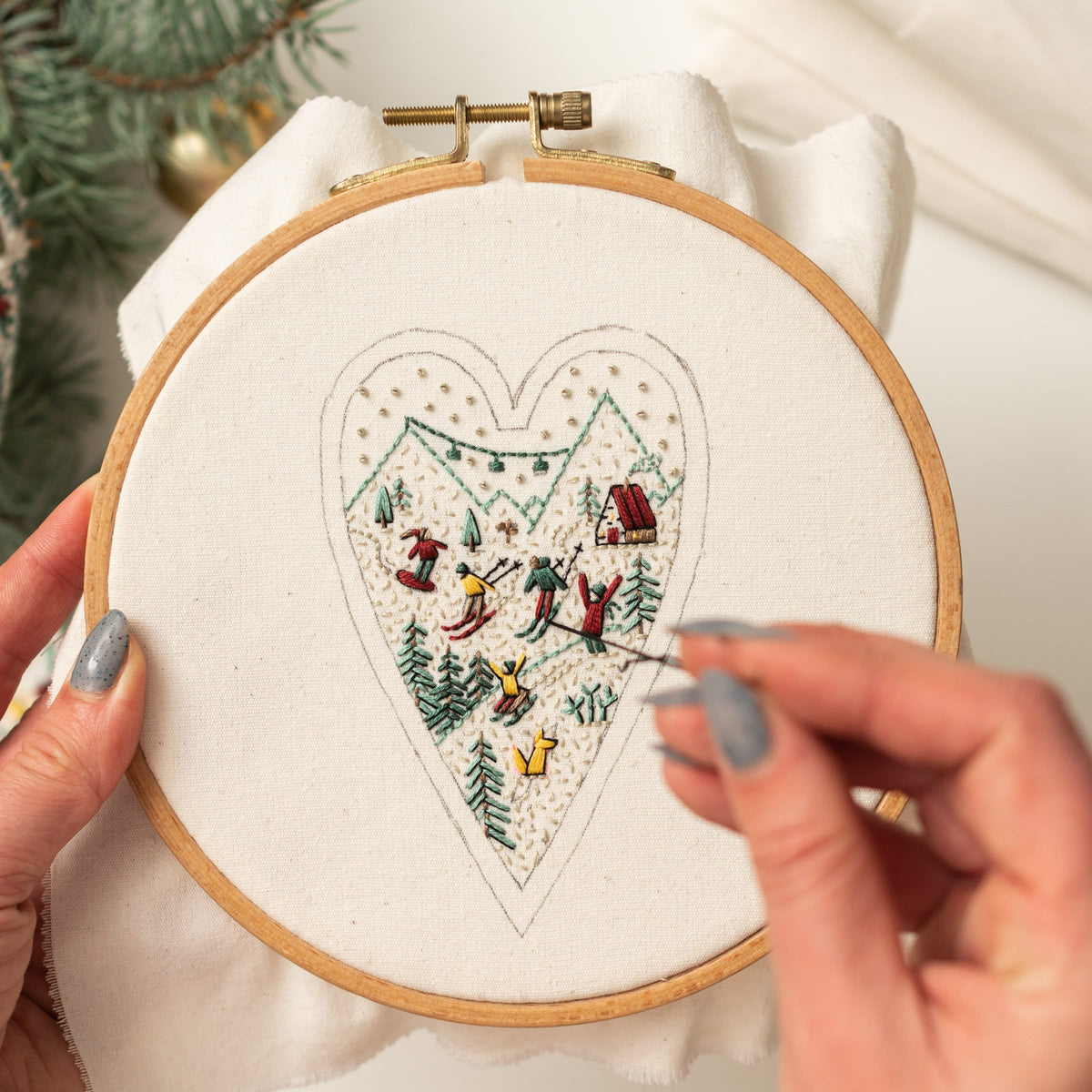 May include: A close-up of a hand embroidering a heart-shaped design on a white fabric stretched in a wooden embroidery hoop. The design depicts a winter scene with skiers and a small house.