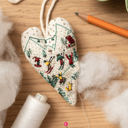 May include: A white heart-shaped ornament with a winter scene embroidered on it. The scene includes skiers, a house, and trees. The ornament is hanging from a string.